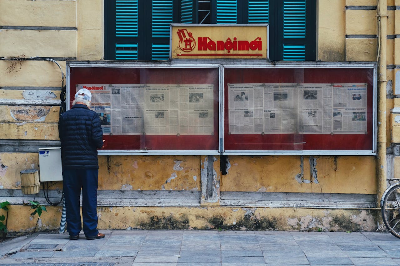 Elderly Man Reading a Newspaper in a Display Case on the Wall of an Old Building